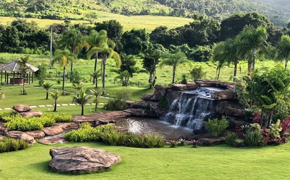 Cachoeira pequena em meio a gramado plano Cachoeira pequena em meio a gramado plano com árvores ao redor e construção ao fundo